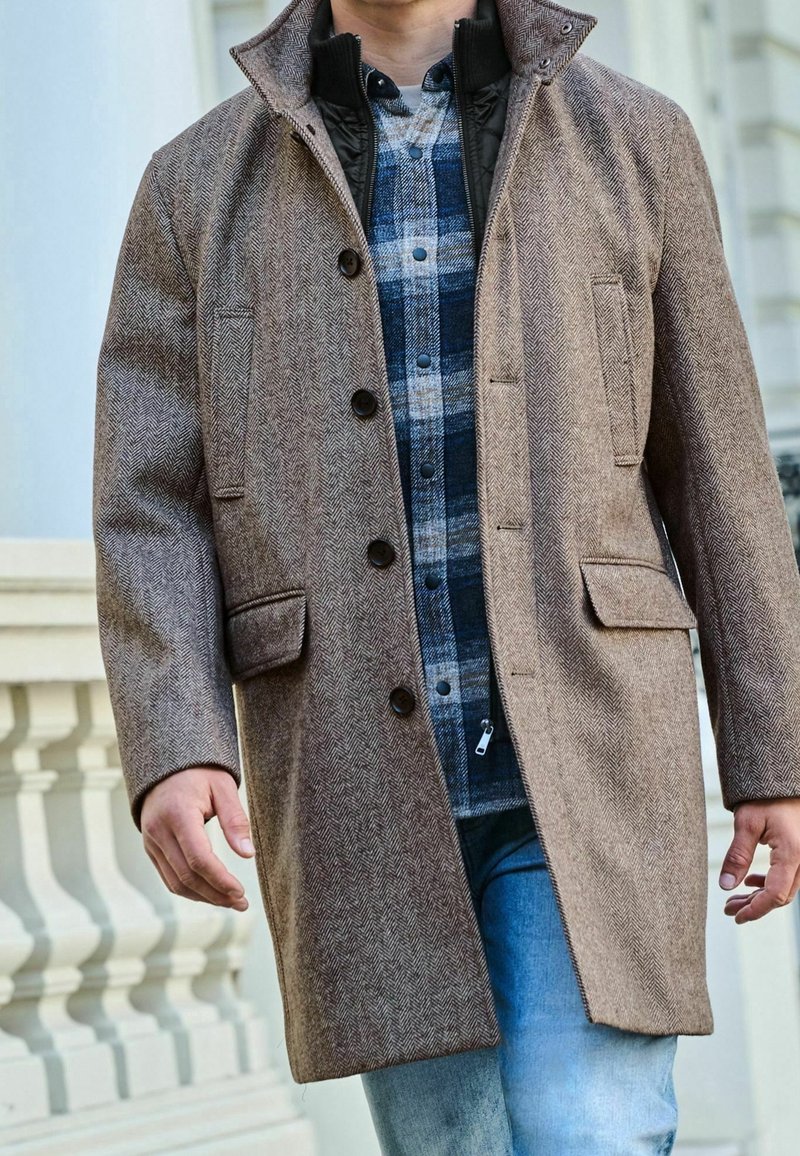 Manteau marron en chevrons avec un col montant, une fermeture à boutons et deux poches avant, porté par-dessus une chemise à carreaux et un jean.