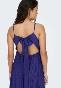 Vestido morado sin espalda con tirantes finos, que presenta un gran lazo atado en el centro y una falda fluida y texturizada.