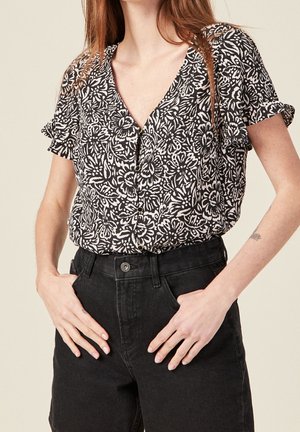 Femme portant un chemisier à manches courtes noir et blanc à motifs floraux boutonné, rentré dans un jean noir taille haute, les mains dans les poches.