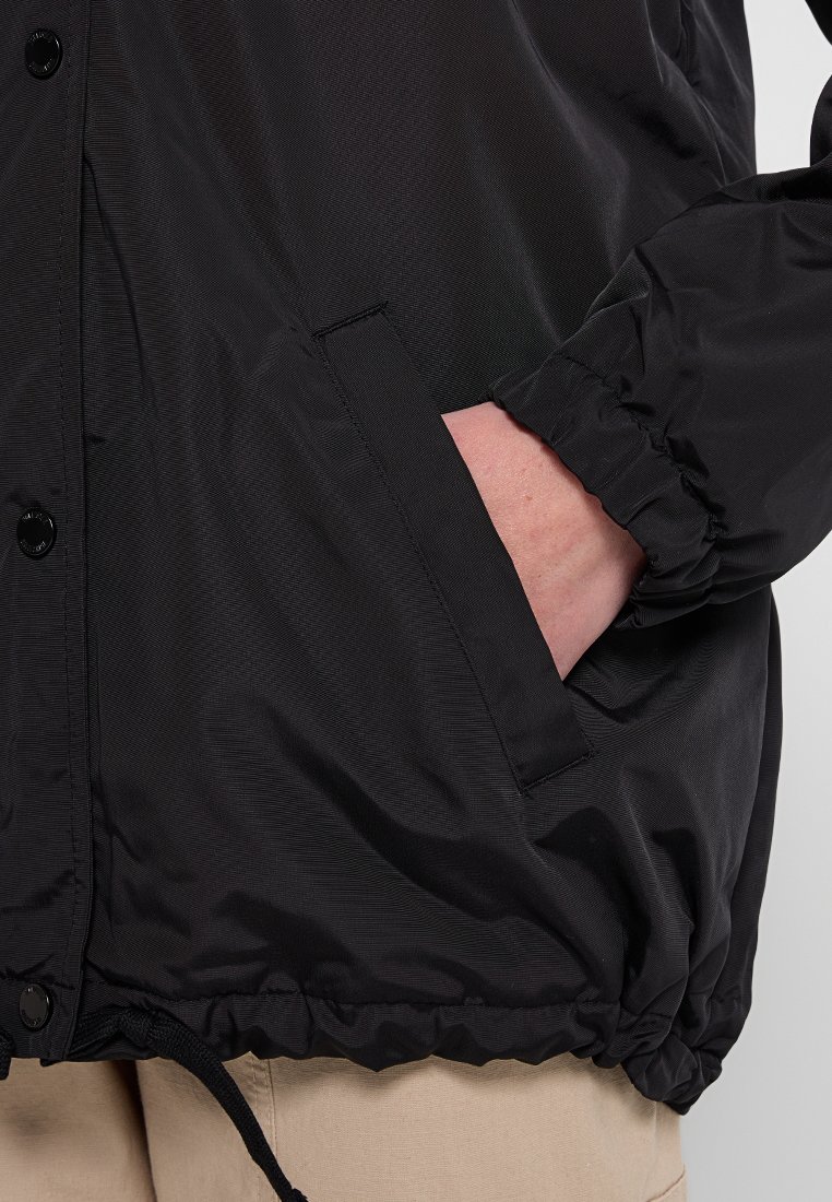 Schwarze Windjacke aus glattem Material, mit einer Reißverschlusstasche, elastischen Bündchen und einem Kordelzugsaum für eine verstellbare Passform.