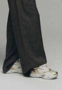 Zapatillas blancas y doradas con materiales texturizados, con forma redondeada y acentos plateados, combinadas con pantalones oscuros y holgados.