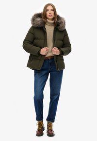 Chaqueta acolchada verde con capucha forrada de piel, suéter de punto beige, vaqueros azules y botas marrones con cordones. Materiales con textura, corte informal.