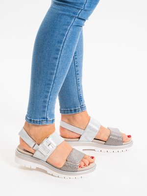 Legs in blue skinny jeans wearing silver open-toe sandals with wide straps, buckle details, and white thick soles on a white background.