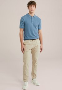 Polo de textura azul claro con mangas cortas, combinado con chinos beige y zapatillas blancas. El modelo se encuentra de pie sobre un fondo neutro.
