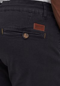 Dark blue trousers with a textured fabric. Features a brown leather logo patch, a single button closure, and a back pocket with yellow stitching.