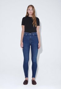 Jeans de denim azul de cintura alta con un ajuste slim y un diseño clásico de cinco bolsillos, combinados con una camisa negra de manga corta y mocasines oscuros.