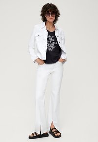 Weiße Jeansjacke, schwarzes Grafik-T-Shirt, weiße Schlaghose mit seitlichen Schlitzen und schwarze Sandalen. Lockige Haare und übergroße Sonnenbrille.