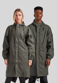 Abrigos largos e impermeables en verde oliva oscuro con puños elásticos, bolsillos con cremallera y capucha ajustable. Textura suave y corte relajado.