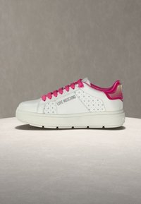 Λευκό αθλητικό παπούτσι Love Moschino με ροζ κορδόνια και ταμπελάκι στη φτέρνα, μικρά γκρι μοτίβα καρδιάς στα πλαϊνά πάνελ, παχύ πάτο σε ουδέτερο φόντο.