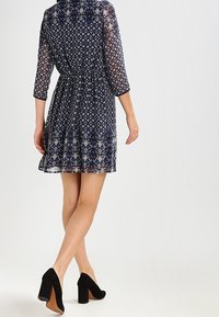 Mörkblå mönstrad klänning med trekvartsärmar, elastisk midja och genomskinligt tyg. Bärs med svarta högklackade skor.