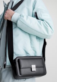 Černá kožená crossbody taška s obdélníkovým tvarem, s přední klopou a stříbrnými detaily. Nošena přes světle modrou zimní bundu.