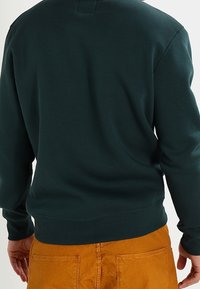Sudadera verde oscura con cuello redondo y puños acanalados, hecha de tela suave, combinada con pantalones de color mostaza. Textura lisa.
