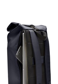 Sac à dos à rabat en tissu bleu marine, avec des bretelles réglables et un profil slim. Comprend des compartiments pour un ordinateur portable et un smartphone.