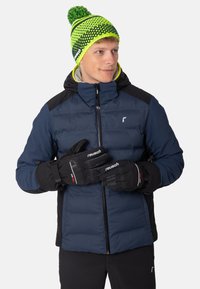 Marineblauwe pufferjas met zwarte accenten, voorzien van een ritssluiting aan de voorkant en een kap. Zwarte handschoenen met rood en wit merklogo. Gebreide muts met groene patronen.