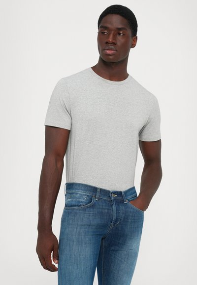 Jeune homme à la peau foncée portant un t-shirt gris clair à manches courtes et un jean bleu, debout avec une main dans la poche.