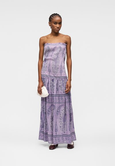 Femme portant une robe longue sans bretelles à motif cachemire violet, tenant une pochette blanche, avec des chaussures bordeaux, debout devant un fond uni.