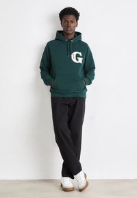 Felpa verde scuro con tasca frontale, caratterizzata da un logo "G" ricamato in bianco. Accoppiata con pantaloni neri e sneaker bianche con suola in gomma.