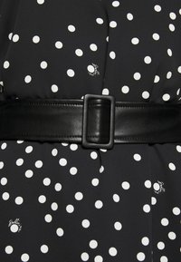 Musta mekko valkoisilla pilkuilla, jossa on leveä musta nahkavyö neliönmuotoisella soljella. Sujuva kangas, jossa on kevyt kiilto ja hillitty brändityyli.