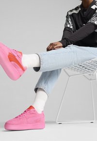 Růžové semišové tenisky s oranžovými akcenty, plochou podrážkou a kulatou špičkou, doplněné o světle modré džíny a bílé ponožky. Jednoduchý design.