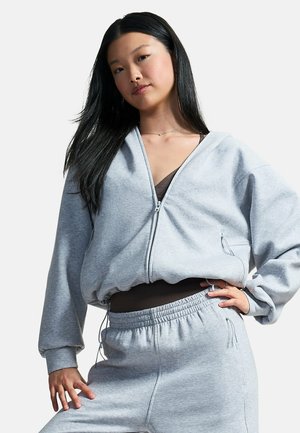 Jonge vrouw met lang zwart haar die een lichtgrijze hoodie met rits en bijpassende joggingbroek draagt tegen een effen achtergrond.