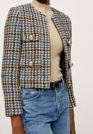 Kobieta nosząca krótką kurtkę w kolorach niebieskim, brązowym i kremowym w wzór pepitki z złotymi guzikami, na beżowym prążkowanym topie na zamek oraz wysokim stanem niebieskich dżinsach.