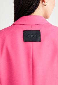 Rozā žakete ar strukturētu apkakli, kurai ir melna birka ar iegravētu vārdu "HUGO" un sarkaniem izšuvumiem.