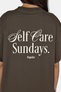 Hnedé bavlnené tričko s voľným strihom. Zadná strana obsahuje biele písmená: "Self Care Sundays." a "Pegador" v jednoduchom písmovom štýle.