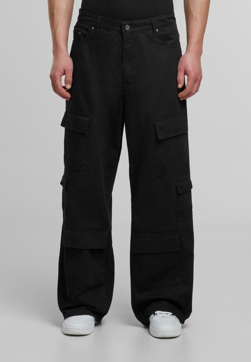 Svarta cargobyxor gjorda av tåligt tyg, med flera fickor, en lös passform och flat front. Bärs med vita sneakers.