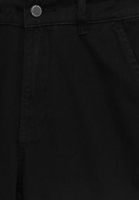 Detailní záběr na černé denimové džíny, ukazující pás, knoflík, kroužek na opasek a švy přední kapsy.