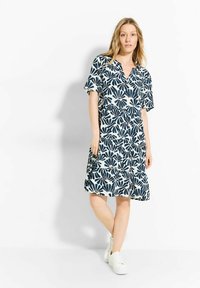 Abito a maniche corte con scollatura a V, realizzato in un tessuto leggero. Presenta un motivo floreale blu navy su sfondo bianco. Il modello indossa sneakers bianche.