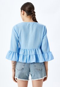 Lichtblauw peplumtop met driekwart mouwen met ruffles, gecombineerd met vervaagde denimshorts. De stof lijkt licht en textuur te hebben.