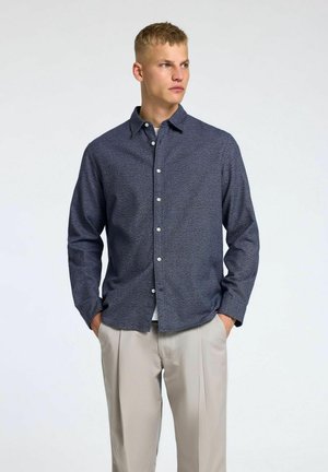 Chemise boutonnée bleu marine à motifs, avec col, manches longues et boutons blancs. Associée à un pantalon beige, soulignant un look smart-casual.