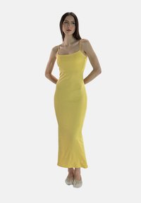 Geel nauwsluitende maxi-jurk met dunne spaghetti bandjes en een gladde, geribbelde textuur, gecombineerd met platte, versierde zilveren schoenen.