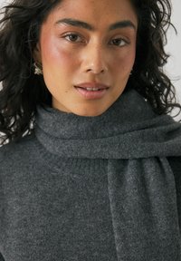 Grijze coltrui met een zachte textuur en een geweven sjaaldetail rond de hals. Heeft een natuurlijke, subtiele make-up en krullend haar.