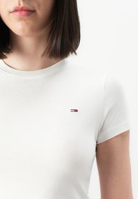 Femeie tânără cu păr de lungime medie, închis la culoare, purtând un tricou simplu alb, cu mânecă scurtă, care are un mic logo roșu, alb și albastru pe piept.