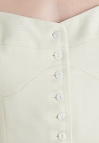 Prima di un abito di stoffa bianco sporco con sei piccoli bottoni rotondi bianchi disposti in una fila verticale e una sottile cucitura curva vicino al collo.