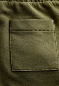 Țesătură din bumbac verde măsliniu cu o textură nervată, având o pungă pătrată coasă pe material. Curea elastică vizibilă în partea de sus.