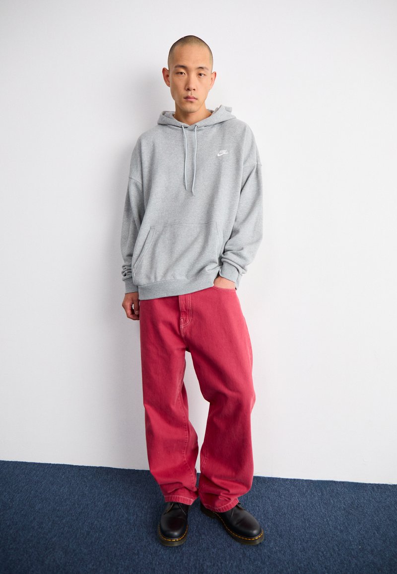 Szary bawełniany hoodie z kieszenią z przodu i logo, w połączeniu z luźnymi czerwonymi dżinsami i czarnymi butami; prosty, casualowy design bez wzorów.