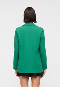 Femme aux cheveux courts et foncés, portant un blazer vert vif ajusté et une jupe noire courte, debout devant un fond blanc uni.