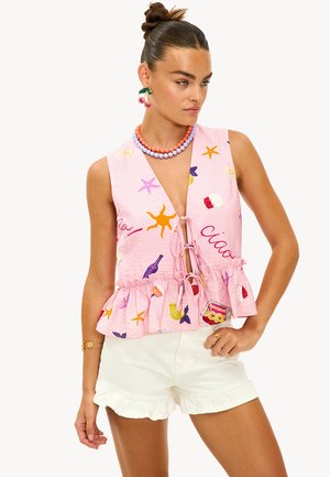 Femme avec des cheveux en chignon portant un haut sans manches rose avec des imprimés ludiques et un short blanc à volants, accessoirisée avec des perles colorées et des boucles d'oreilles en forme de cerise.