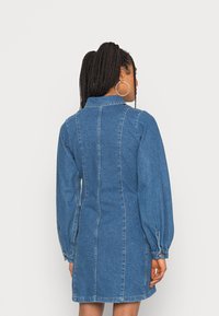 Vestido de camurça azul com cintura ajustada, mangas bufantes longas e detalhes na costura das costas. Apresenta um colarinho e punhos com botões.