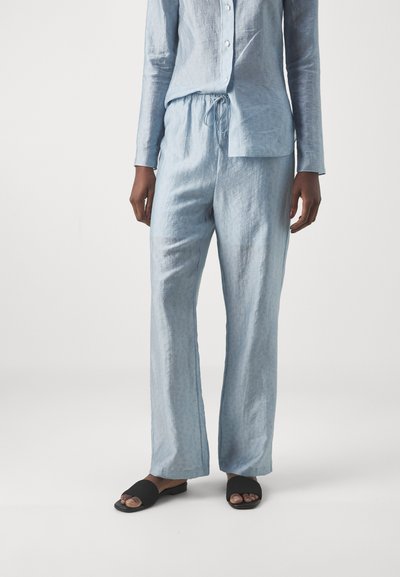 Chemise en lin bleu clair à manches longues et boutons, associée à un pantalon en lin bleu clair assorti. Des sandales noires à enfiler complètent la tenue.