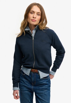 Marineblå sweatshirt med lynlås og raglanærmer, tekstureret stof og broderet logo. Lag-på-lag med en let stribet skjorte under.