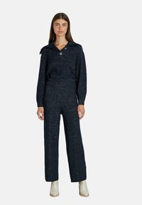 Frau in einem dunkel karrierten Strickpullover und passenden weit geschnittenen Hosen, kombiniert mit weißen Stiefeletten, stehend vor einem schlichten Hintergrund.