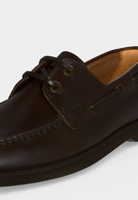 Chaussure bateau en cuir marron foncé avec lacets noirs, coutures visibles et doublure intérieure couleur beige sur un fond clair uni.