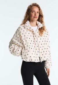 Nő hosszú, hullámos hajjal, fehér, fekete pöttyös steppelt dzsekit, fehér felsőt és fekete leggingset visel, sima világos háttér előtt áll.