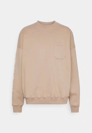 Beige sweater met ronde hals en lange mouwen, een borstzak aan de voorkant, geribde manchetten en zoom, en wit bedrukt tekst gedeeltelijk zichtbaar op de linkermouw.
