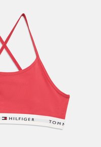 Sarkans sporta krūšturis ar gludu tekstūru, ar šķērsām saslēgtām lencītēm un baltu joslu ar melniem burtiem apakšā.