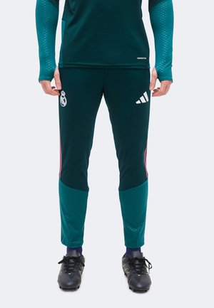 Dolna część ciała osoby ubranej w ciemnozielone spodnie treningowe Real Madrid z logo Adidas oraz czarne korki piłkarskie na białym tle.