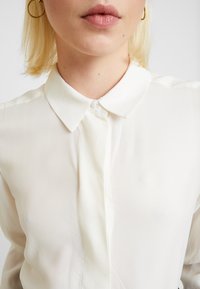 Bílá blůza s límečkem a jemnou diagonální pruhovanou texturou. Obsahuje knoflíkový uzávěr u krku a moderní, jednoduchý design.